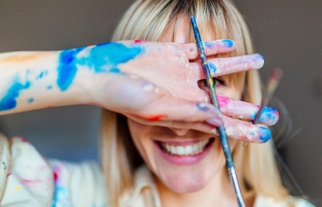 Woman Covering Face with Paint Covered Hand Holding a Paintbrush.png